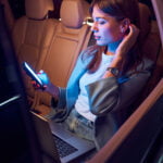 Female blogger with notebook on her laps sitting on back seat in automobile and holding cellphone